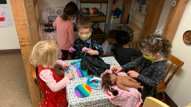 In the foreground, children play with dolls and pretend household items. In the background, children play in a pretend kitchen area.