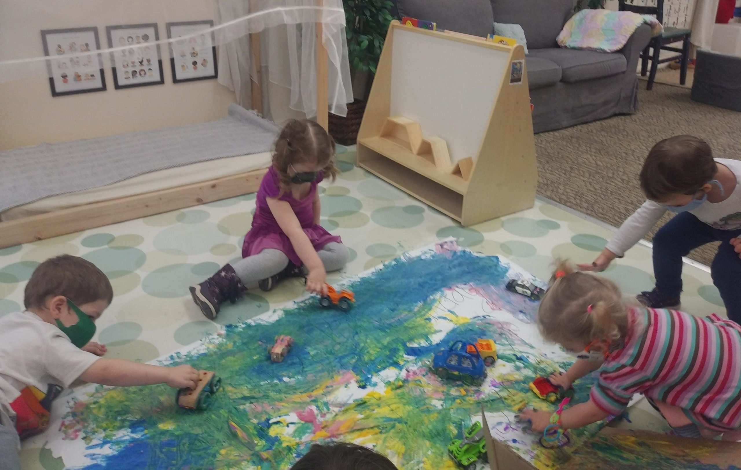 Children drive toy cars through blue and yellow paint, making the color green. In the background is a quiet space and a shelf with books and a white board.