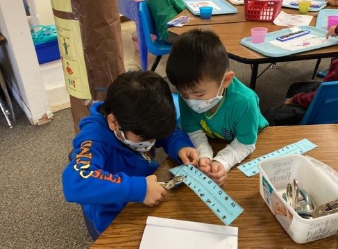 Two boys work together to use a hole punch on a strip of paper with numbers on it. In the background, children explore with watercolor paints.