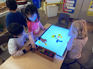 Three young children explore transparent shapes on top of a lighted board in their classroom.